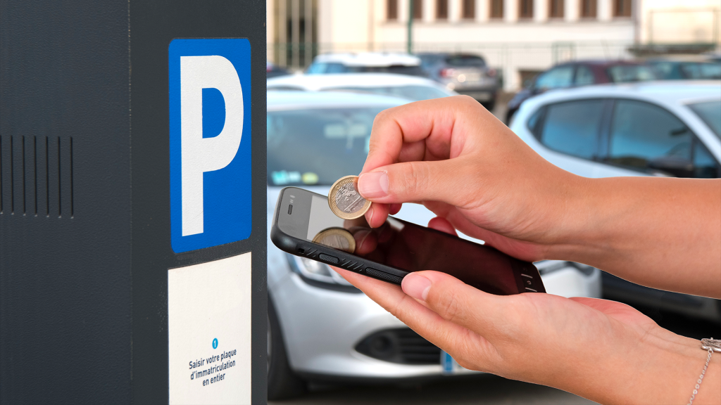 Une personne tient une pièce de deux euros au-dessus d’un smartphone, devant un horodateur de stationnement. En arrière-plan, on voit des voitures stationnées dans une rue. Sur l’horodateur, un grand "P" blanc sur fond bleu indique qu'il s'agit d'une borne de stationnement payant.